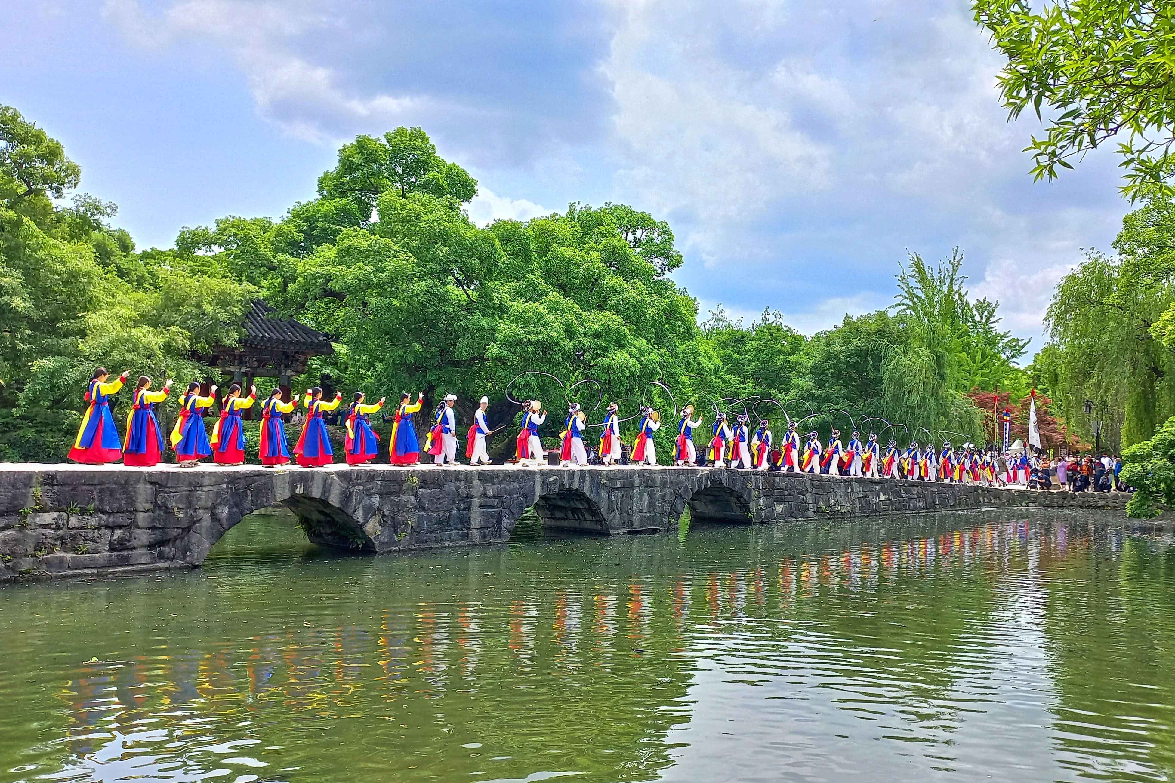 Performers in colorful dress lined along a stone bridge over the pond at Gwanghalluwon Garden in Namwon.